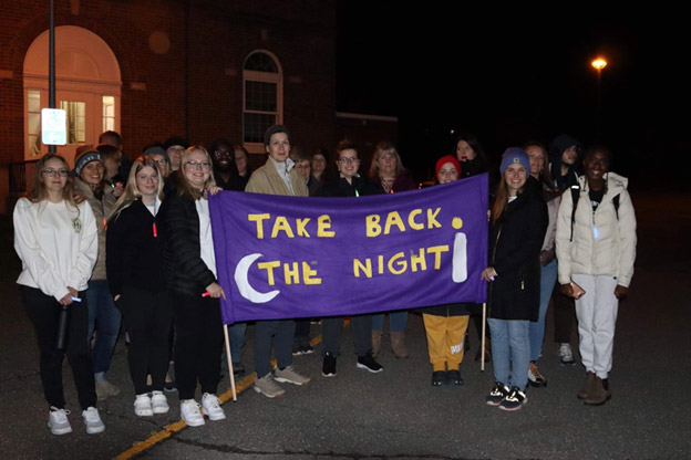 Members of the Student Organization of Social Work during an event smile for the camera with a purple banner that reads "Take Back the Night"