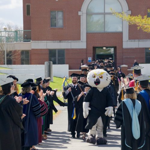 Photo of UMPI graduating students marching down the campus walkway as faculty and staff cheer them on.