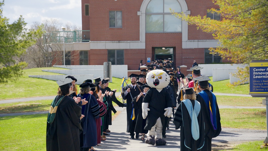Photo of UMPI graduating students marching down the campus walkway as faculty and staff cheer them on.